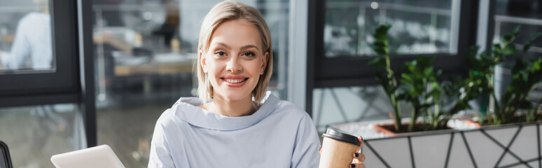 Positive businesswoman with coffee to go and digital tablet looking at camera in office, banner.