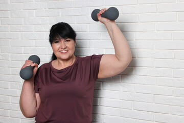 Happy overweight mature woman doing exercise with dumbbells near white brick wall