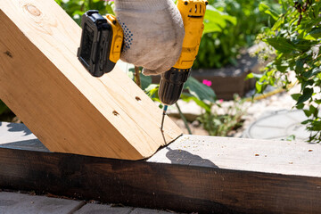 A carpenter works with a yellow electric screwdriver, twists a screw into a wooden plank beam