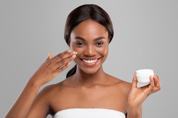 Glad young black woman with perfect skin applying moisturizing anti-aging cream on her face and hold jar