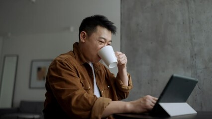 Smiling Asian man watching something on tablet and drinking coffee at home - Powered by Adobe