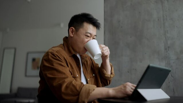 Smiling Asian man watching something on tablet and drinking coffee at home