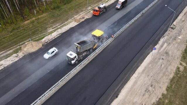 The yellow paver is moving slowly down the highway. The paver distributes and compacts the hot asphalt paving. Road workers operate the equipment and make road repairs. Annual maintenance of freeway