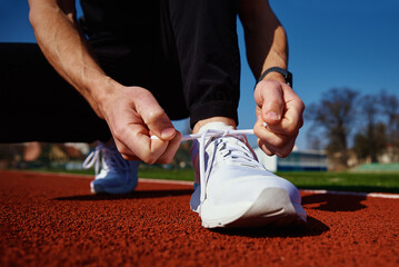 Male runner in white sneakers get ready for run at stadium track. Male hands tying on sport...