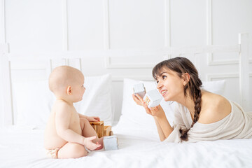 a young mother plays with a baby in diapers with wooden toys on a bright bed at home and smiles