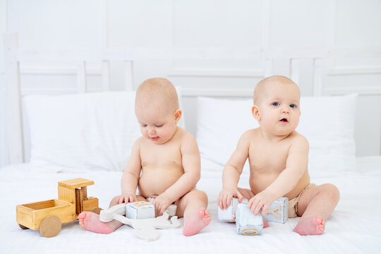 two twin babies girls of six months in diapers on a white cotton bed in a bodysuit on a bed at home playing with wooden toys and smiling