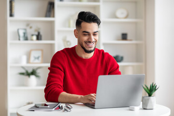 Cheerful attractive millennial arabic businessman with beard in red clothes typing on laptop in home office