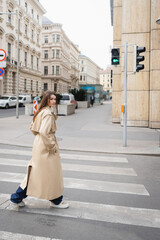 full length of woman in trench coat passing crosswalk in european city.