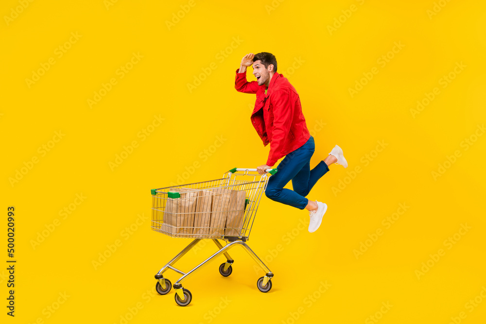 Poster full length body size view of attractive cheerful guy jumping carrying cart looking search goods iso
