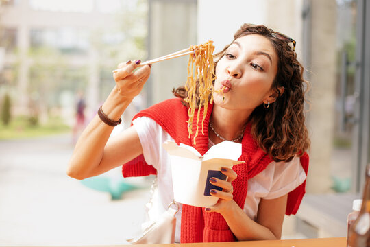A happy Asian girl holds Chinese chopsticks and eats wok noodles from a take-away paper box. Street and fast food concept