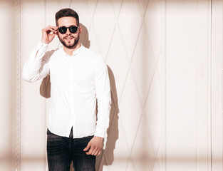 Handsome confident  model. Sexy stylish man dressed in white shirt and jeans. Fashion hipster male posing near white wall in studio interior at sunny day. Shadow from window. In sunglasses
