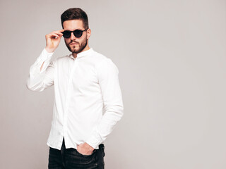 Fototapeta premium Portrait of handsome confident model. Sexy stylish man dressed in white shirt and jeans. Fashion hipster male posing in studio on grey background. In sunglasses. Isolated
