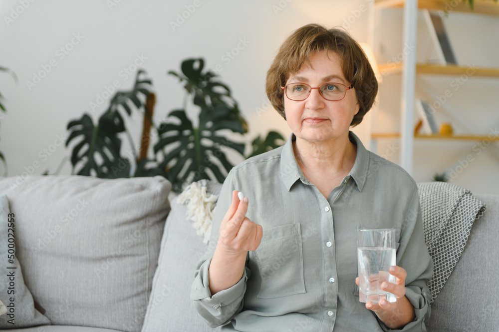 Wall mural age, medicine, healthcare and people concept - senior woman with glass of water taking pills at home