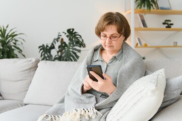 older mature adult woman using smartphone sitting on couch at home. Cheerful grandmother with eyeglasses surfing internet with her mobile phone. Technology concept