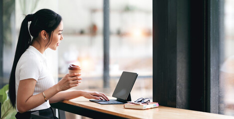 Young asian businesswoman holding cup and using digital tablet while sitting in co-workspace.