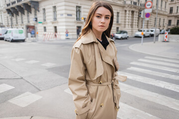 young woman in stylish trench coat standing with hands in pockets on street of european city.