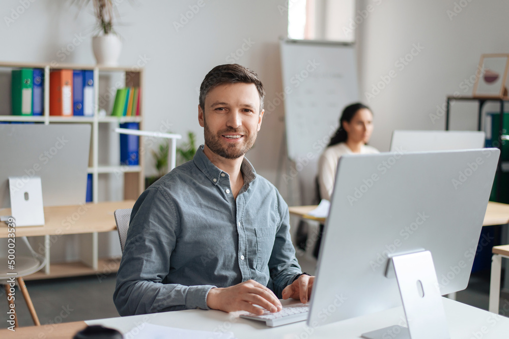 Poster Handsome cheerful man sitting at desk, using modern computer in open space office