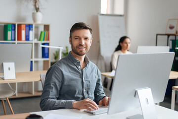 Handsome cheerful man sitting at desk, using modern computer in open space office