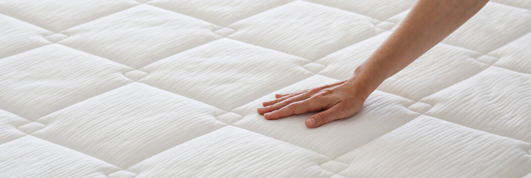 Cropped shot of young woman's hand testing white orthopedic matress on firmness. Female pressing hypoallergenic foam mattress surface to check its softness. Close up, copy space, top view, background.