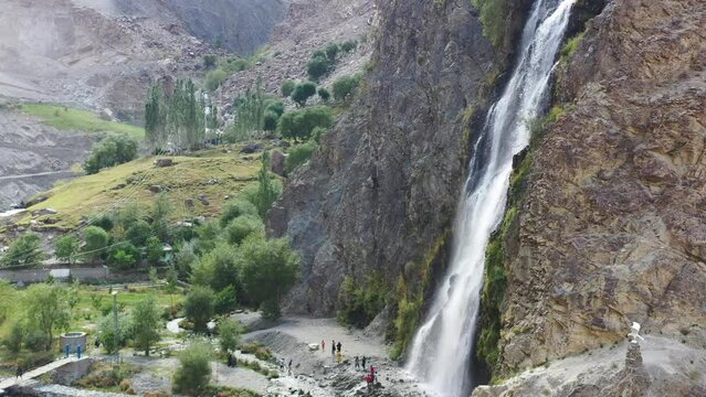 aerial drone panning across left at Manthokha Waterfall in Skardu Pakistan as tourists below are surrounded by mountains taking photos on a sunny summer day