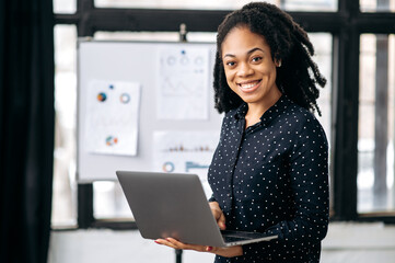 Portrait pretty African American business woman, financial manager or mentor, broker, marketer, stand in office, against a white board with charts, holds an open laptop in hands, looks at camera,smile
