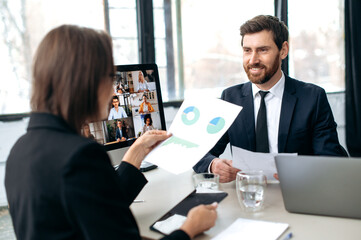 Two influential successful business colleagues, working together in modern office, study financial documents, talking with partners by online video conference by computer, discuss business strategy