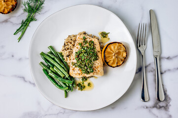 Top view of halibut fish with chimichurri sauce, green beans, lemon on a white plate on marble table - obrazy, fototapety, plakaty