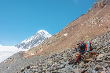 Tourist with camera sitting on stone in high mountains in sunny day. Scenic alpine landscape with man in bright sun against snow mountain peak and rocks in sunlight. Backpacker among snow mountains.