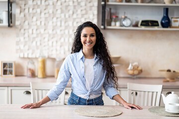 Portrait of excited latin housewife standing near kitchen table, looking at camera and smiling, free space