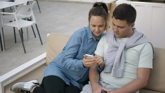 Portrait of young caucasian woman and man wearing casual clothes. Beautiful young girl showing the screen of smartphone sitting on sofa with her boyfriend on hotel terrace background. High angle view