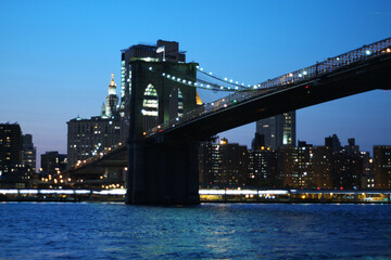Famous Brooklyn Bridge and the NYC skyline behind with lights on in the evening