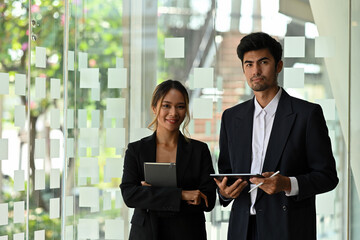 A portrait of Asian businesspeople standing in a workplace holding tablets and looking at the camera, for business and technology concept.