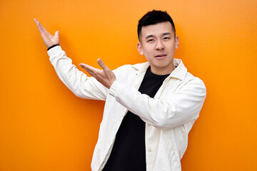 Young Chinese man in white shirt on orange background isolated smiling and looking at the camera pointing with both hands to the side. High quality photo