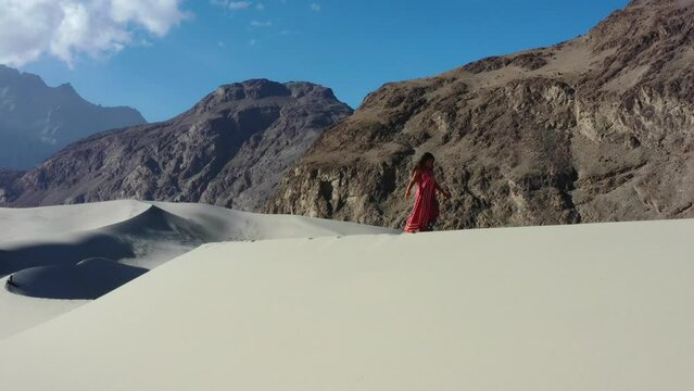 aerial drone tracking a young asian female tourists with a red dress walking the top of sand dunes in the Cold Desert of Skardu Pakistan with the rugged mountain range in the background on a sunny day