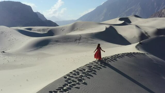 aerial drone of a young asian female tourists in a red dress standing the top of sand dunes in the Cold Desert of Skardu Pakistan with the rugged mountain range in the background on a sunny day