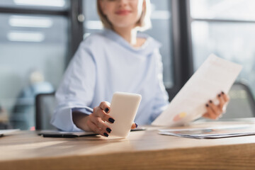 Cropped view of blurred businesswoman using smartphone near papers in office.