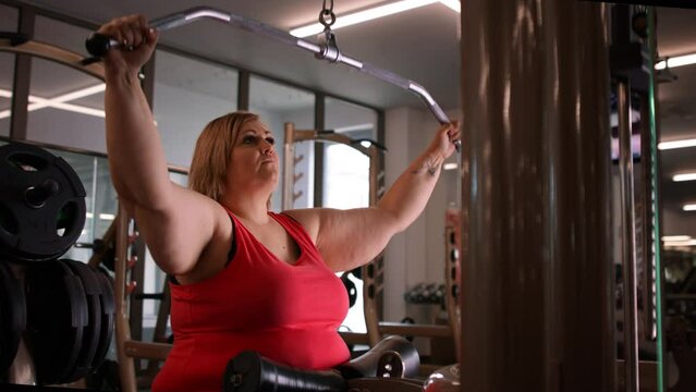 Fat woman working out on a lat pulldown machine indoors in gym