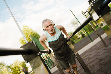 Gray mature man listening music while working out on parallel bars