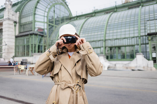 happy young woman in beige trench coat and baseball cap taking photo on vintage camera.
