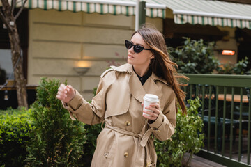 young woman in trendy sunglasses and trench coat holding paper cup.