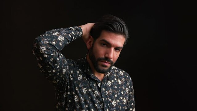 Close up studio portrait of handsome seductive confident intriguing masculine man in floral long-sleeved shirt touching his shiny healthy hair. Black background. High quality 4k footage
