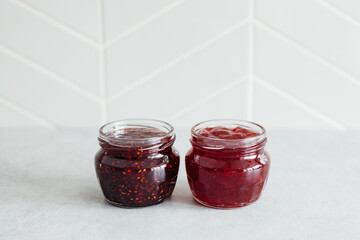Two open glass jars with red strawberry and raspberry jam on grey table against gray tile kitchen...