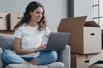 Hispanic girl sitting with laptop among cardboard boxes and smiling. Relocation and online work.