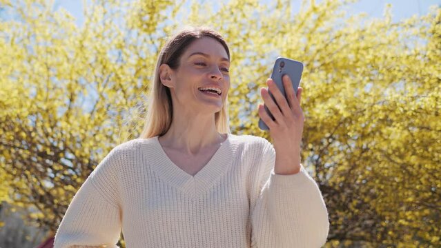 Beautiful woman in a white sweater using mobile phone having online video call chat in the park near a tree with yellow leaves on a warm sunny day. Female lady making conference chat spend free time.