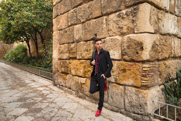 Obraz na płótnie Canvas Young Spanish man, wearing black shirt, jacket and pants, with black scarf with red polka dots, red dancing shoes, posing next to a stone wall. Flamenco concept, art, dance, culture, tradition.