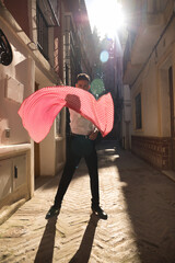 Young Spanish man in white shirt and black dance pants and shoes, dancing flamenco with a red handkerchief with white polka dots in the street, under the sun rays. Concept dance, culture, tradition.