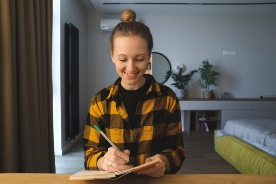 young caucasian woman is smiling, sitting at the desk in her bedroom at home, writing notes, making great plans in her notebook, holding a pen, looking satisfied and happy,wearing checked yellow shirt