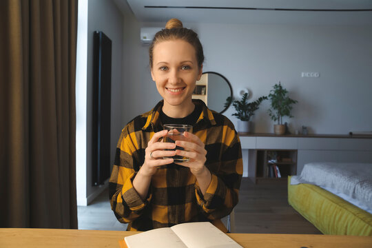 young caucasian woman is sitting at the desk in her bedroom at home, writing down in her notebook, holding glass with water, wearing checked yellow shirt, hair bun, looking at camera, cozy interior