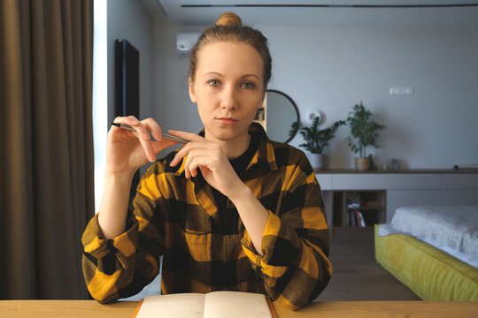 young caucasian woman is sitting at the desk in her bedroom at home, writing notes, making plans in her notebook, holding a pen, wearing checked yellow shirt, hair bun, cozy interior