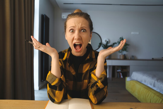 young caucasian woman is sitting at the desk in her bedroom at home, writing notes, surprised or shocked, being struck by the negative news, holding a pen, facial expression concept